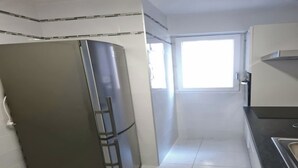 Ett kök med ett kylskåp i rostfritt stål, vita kakelväggar, ett fönster och en svart bänkskiva.