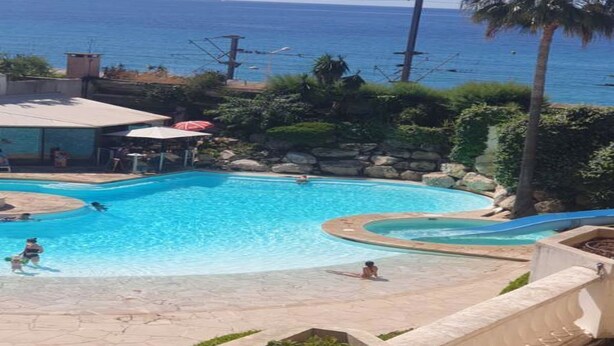 En pool med en vattenrutschkana, omgiven av grönska och med utsikt över havet.
