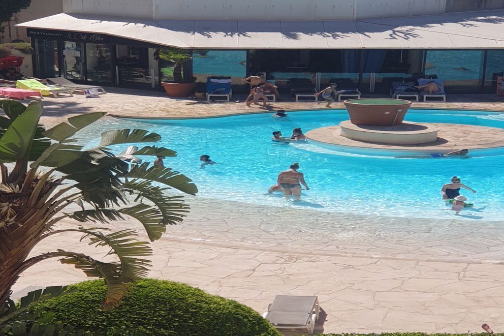 A pool area with people swimming and lounging, surrounded by palm trees and a restaurant in the background.
