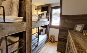 Ein kleines, rustikal eingerichtetes Zimmer mit Etagenbetten, einem kleinen Hocker und einem Holztisch mit einem gerahmten Bild.