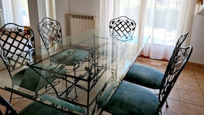 Een eetgedeelte met een tafel met glazen blad en metalen stoelen, een radiator en uitzicht door dunne gordijnen.