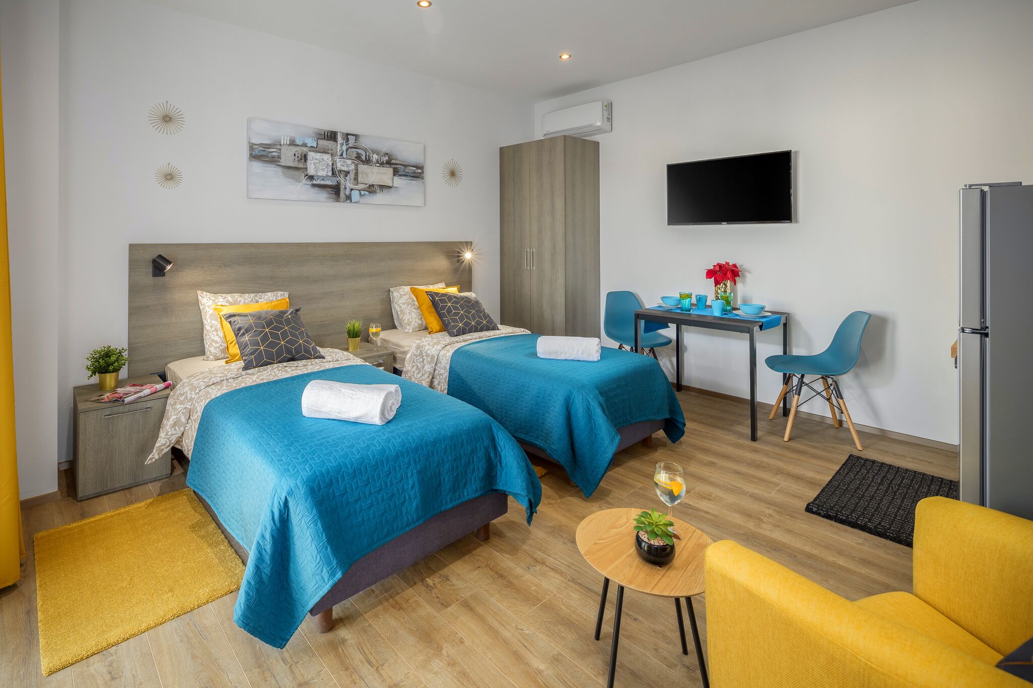 A hotel room with two beds, a desk, a chair, a TV, and a refrigerator.
