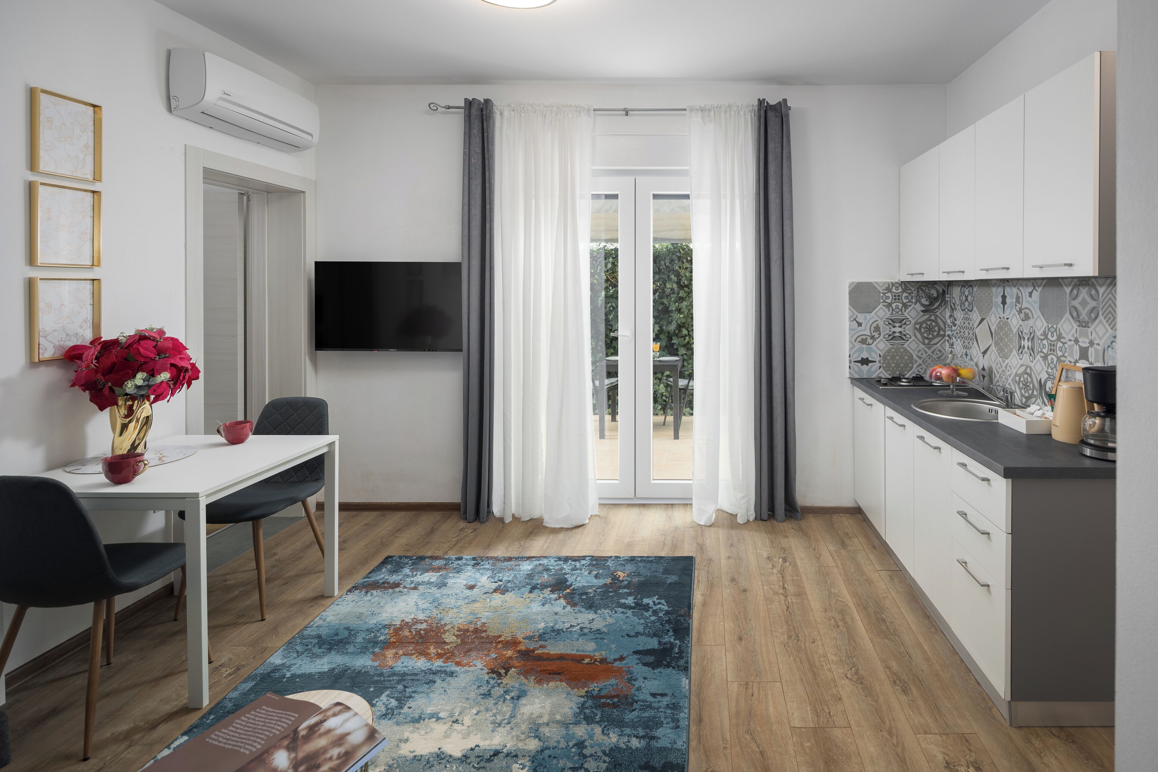 Eine moderne Küche mit Flachbildfernseher, einem Essbereich mit Tisch und Stühlen sowie Blick ins Freie durch eine Glastür.