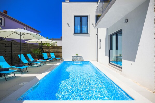A swimming pool with blue tiles, surrounded by lounge chairs and umbrellas, and a modern building with a large window.