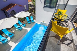 A rooftop pool with blue lounge chairs and yellow chairs, a small table with a plant, and a glass balustrade.