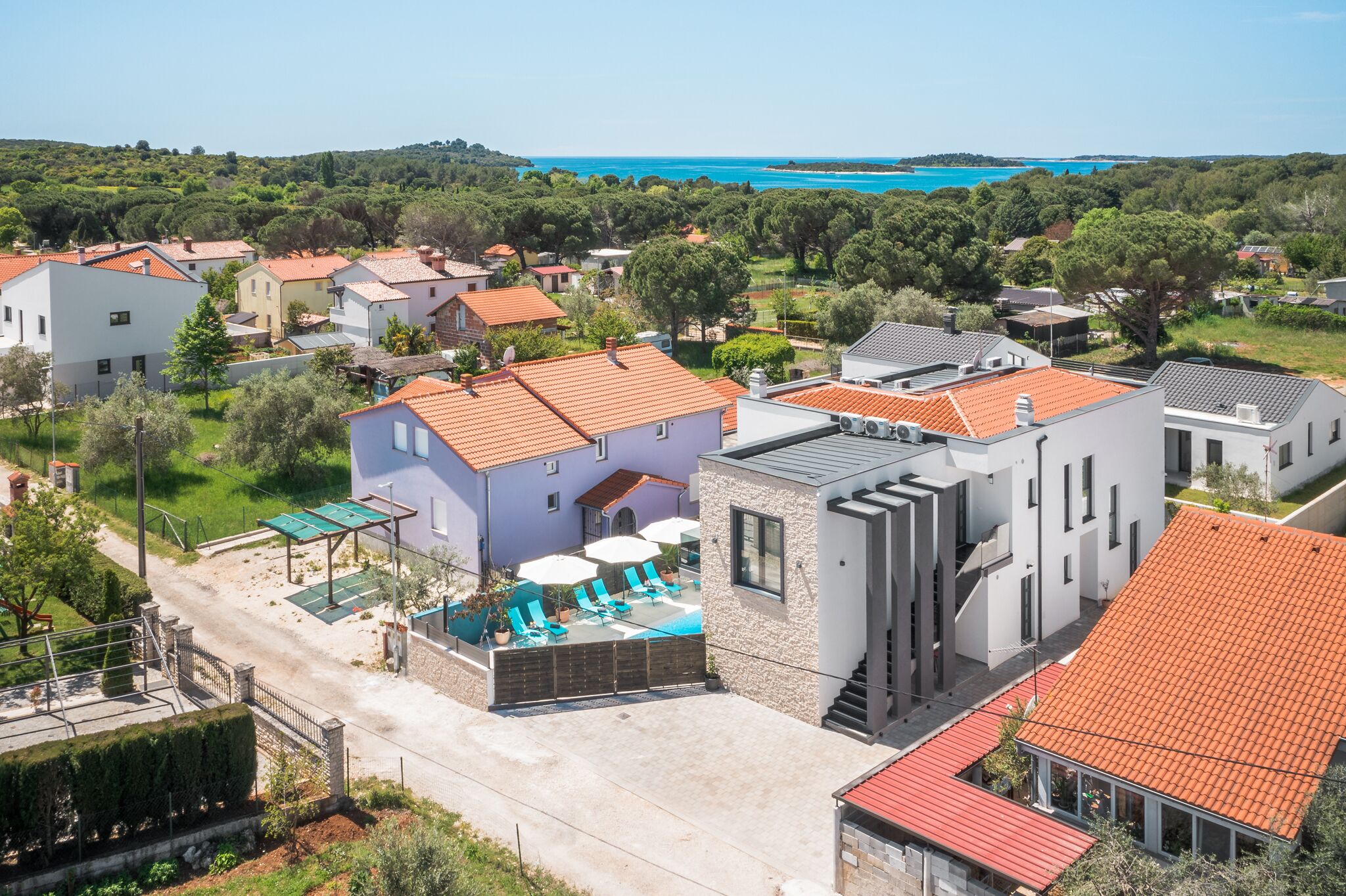 A residential area with a swimming pool, houses with red-tiled roofs, and a view of the sea.