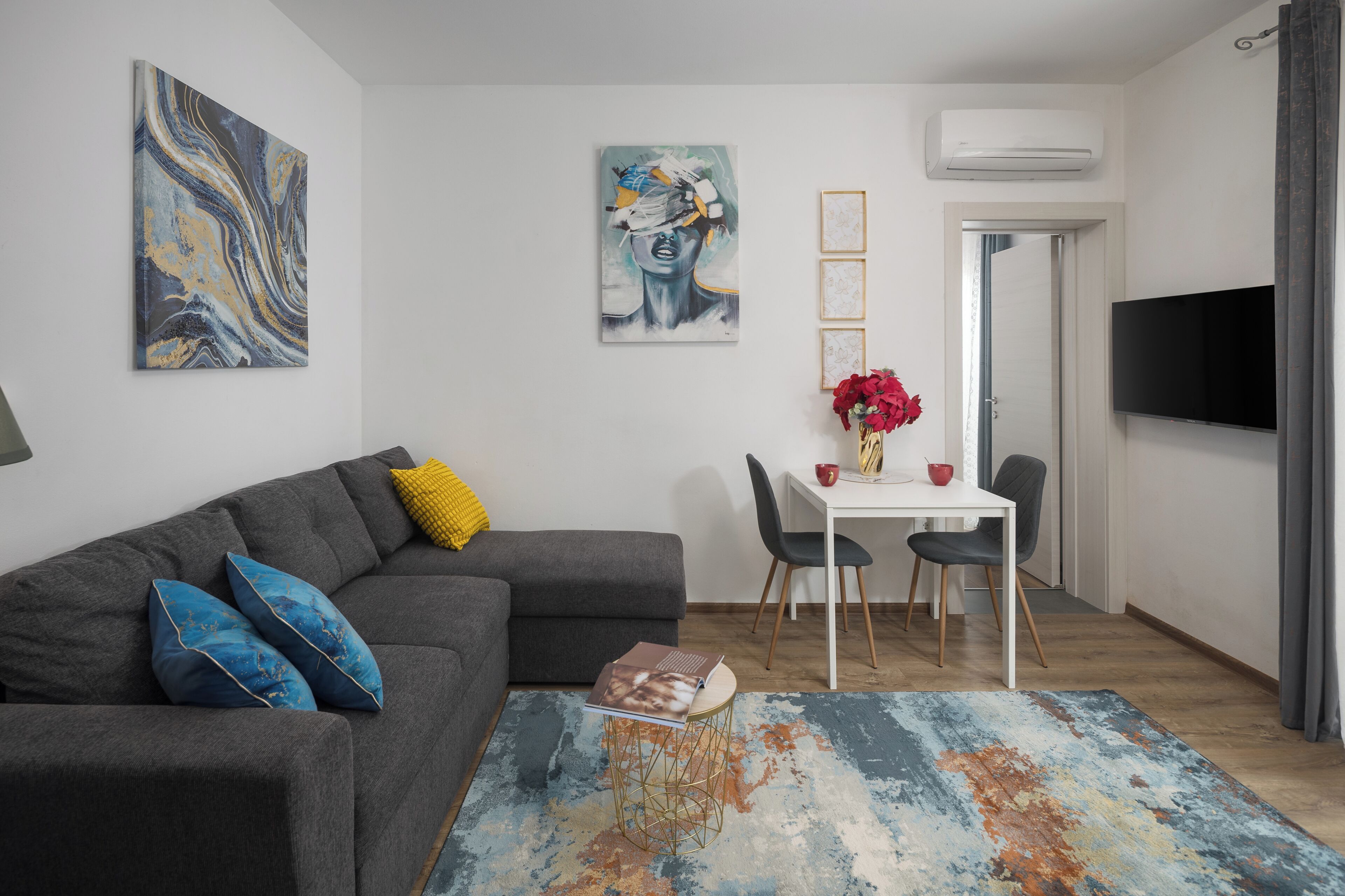 Ein modernes Wohnzimmer mit einem grauen Sofa, einem Couchtisch und einem Essbereich mit weißem Tisch und Stühlen. An den Wänden befinden sich Gemälde und ein Flachbildfernseher ist über dem Esstisch angebracht.