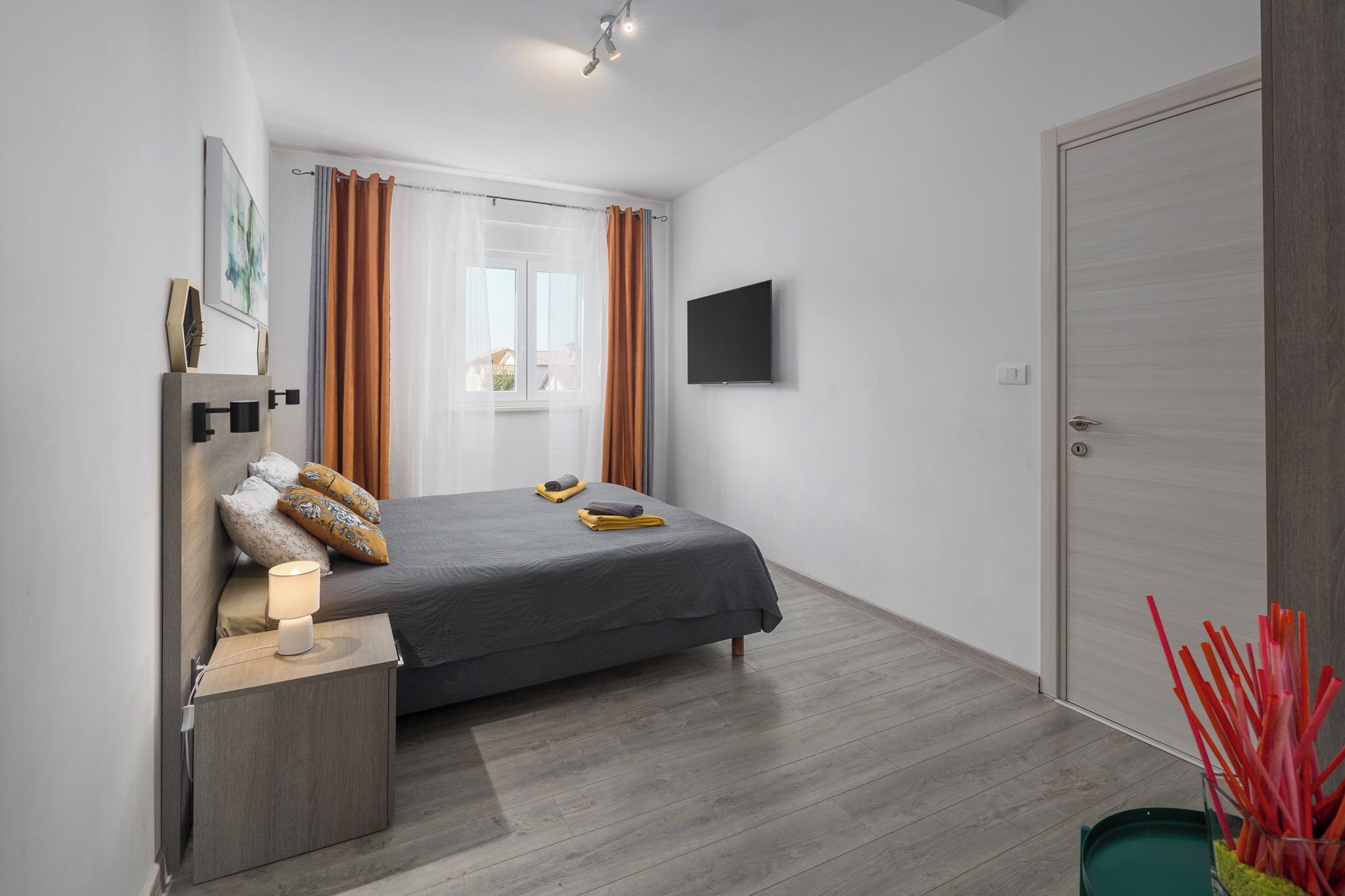 Un dormitorio moderno con cama, mesita de noche, lámpara, cortinas, televisor y puerta.