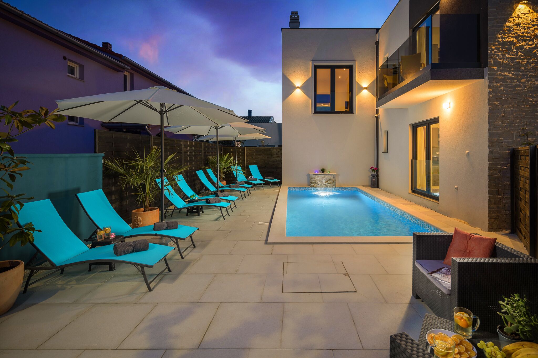 A modern outdoor pool area with lounge chairs and umbrellas, a small pool, and a building with a balcony.