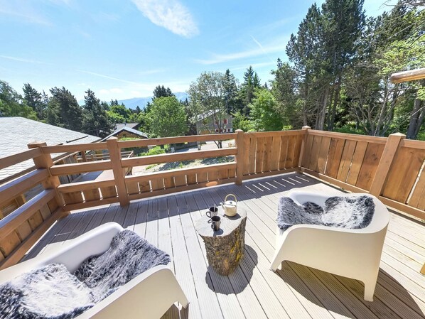 A wooden deck with a wooden fence, two white chairs with fur throws, a small table, and a view of trees and buildings.