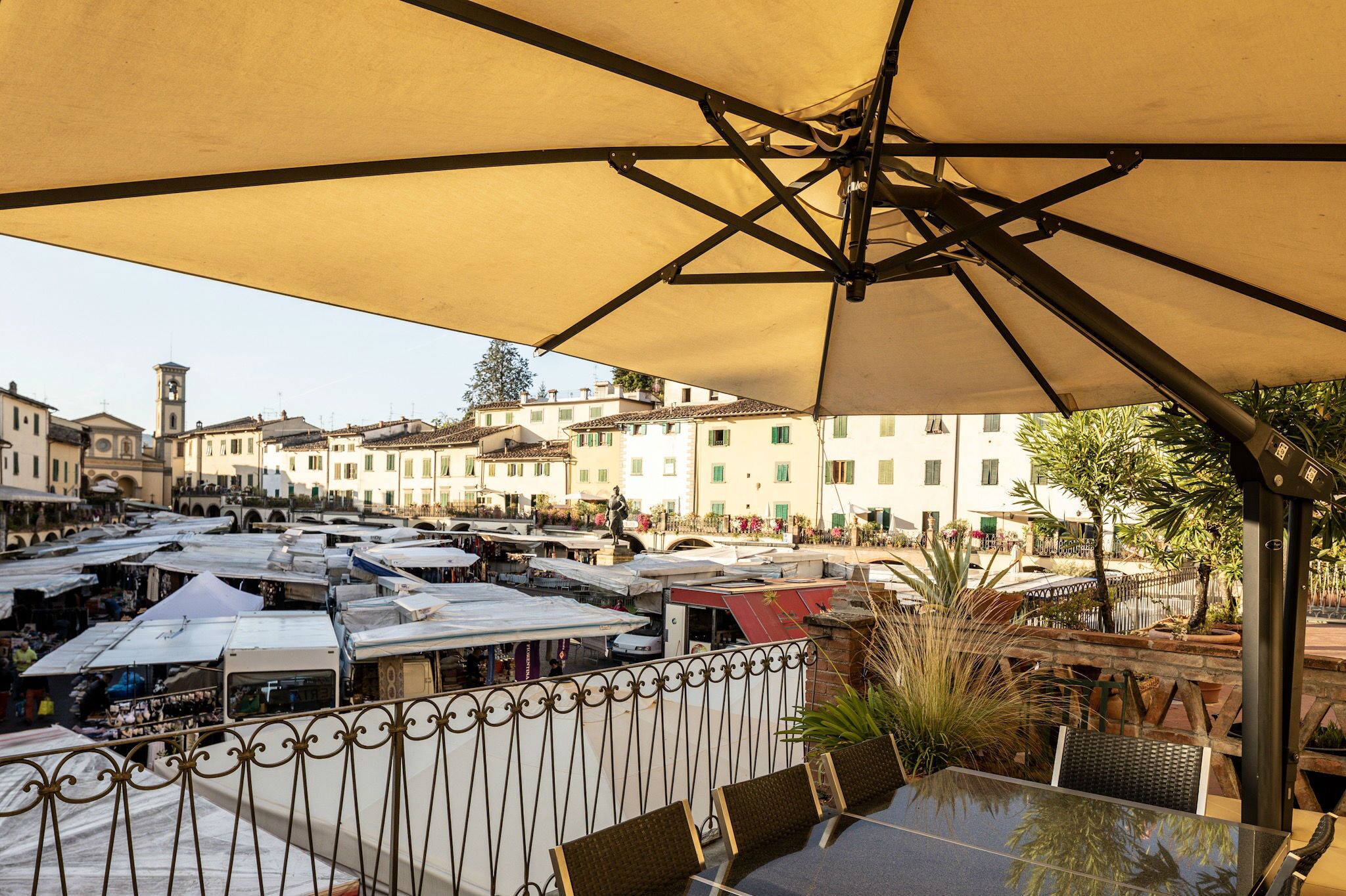 An outdoor seating area with tables and umbrellas, overlooking a bustling market street with buildings and shops.