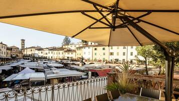 An outdoor seating area with tables and umbrellas, overlooking a bustling market street with buildings and shops.