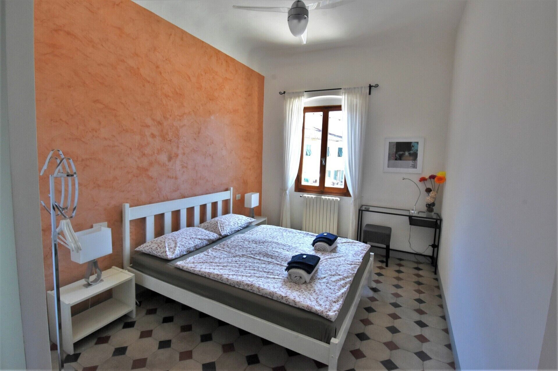 A bedroom with a bed, a nightstand, a radiator, a window with curtains, a desk with a chair, and a wall-mounted picture.