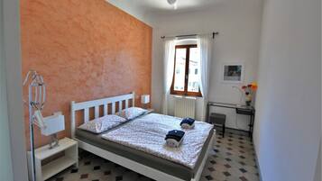 A bedroom with a bed, a nightstand, a radiator, a window with curtains, a desk with a chair, and a wall-mounted picture.