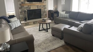 A living room with a stone fireplace, a flat-screen TV mounted above it, a grey sofa, a round coffee table, and a lamp. - Valley View Lakehouse - Fully updated, dog friendly and beautiful lake view! (Mayville, Chautauqua Lake)