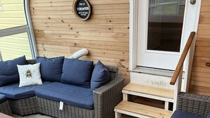 Un espacio exterior acogedor con un sofá de mimbre y un banco de madera, una mesita y un letrero que indica que los muebles se venden al barril.