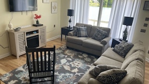Un salon comprenant un canapé, un fauteuil, un téléviseur à écran plat, une table d’appoint et une fenêtre avec des rideaux.