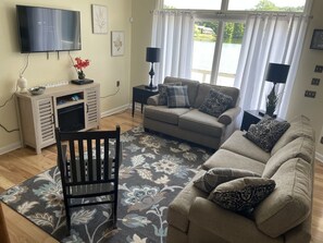 Un salon comprenant un canapé, un fauteuil, un téléviseur à écran plat, une table d’appoint et une fenêtre avec des rideaux.