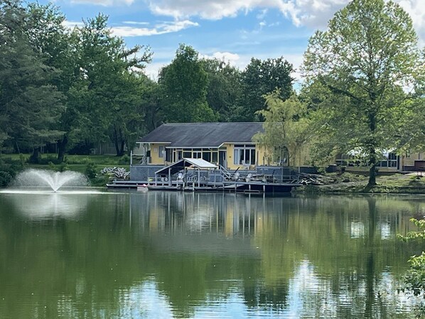 A lakeside house with a dock, a fountain, and surrounding trees.