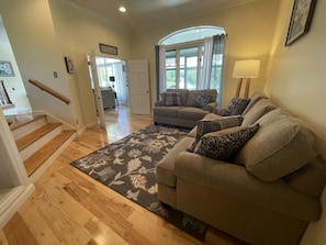 Una sala de estar con un sofá beige, alfombra estampada, piso de madera y una escalera.