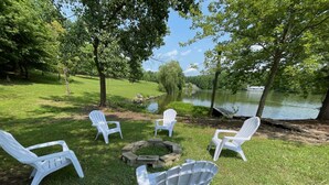 Un espace extérieur verdoyant, avec des chaises Adirondack blanches disposées autour d’un foyer en pierre.