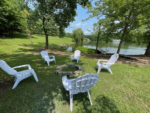 Un espace extérieur verdoyant, avec des chaises Adirondack blanches disposées autour d’un foyer en pierre.