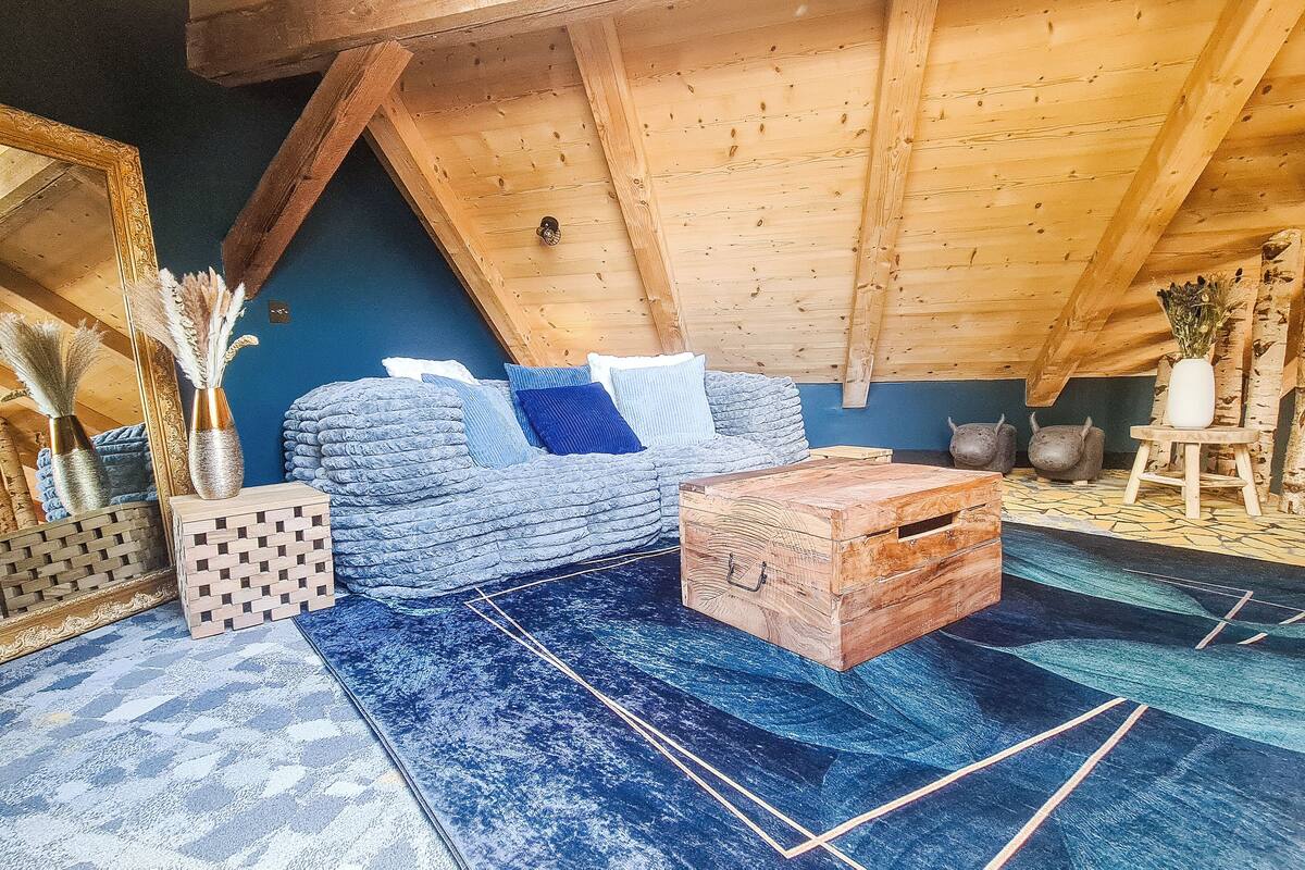 A cozy attic room with a wooden ceiling, a blue sofa, a woven side table, and a patterned rug.