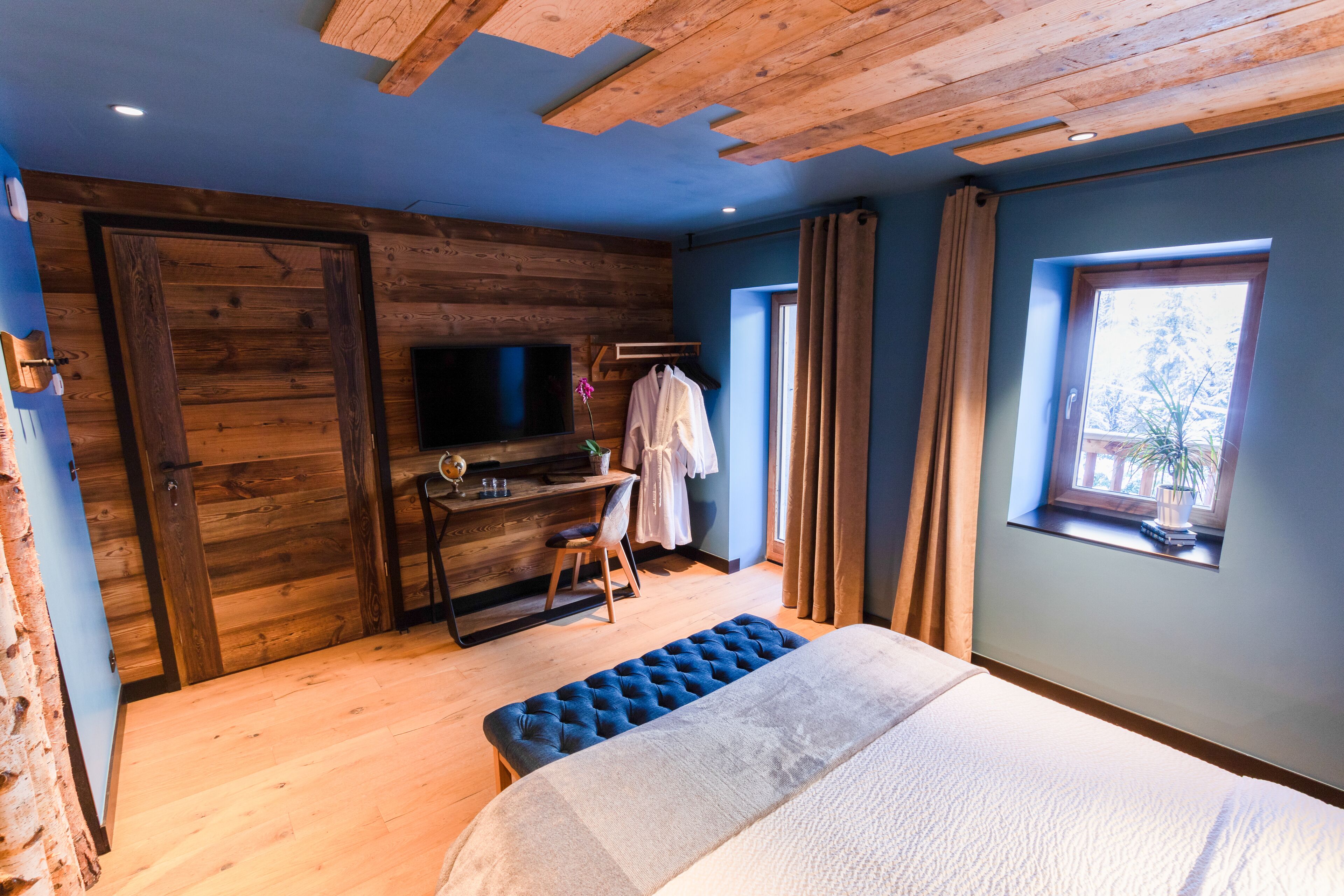 A bedroom with a wooden ceiling, a large bed, a desk with a chair, a TV, and a window with curtains.