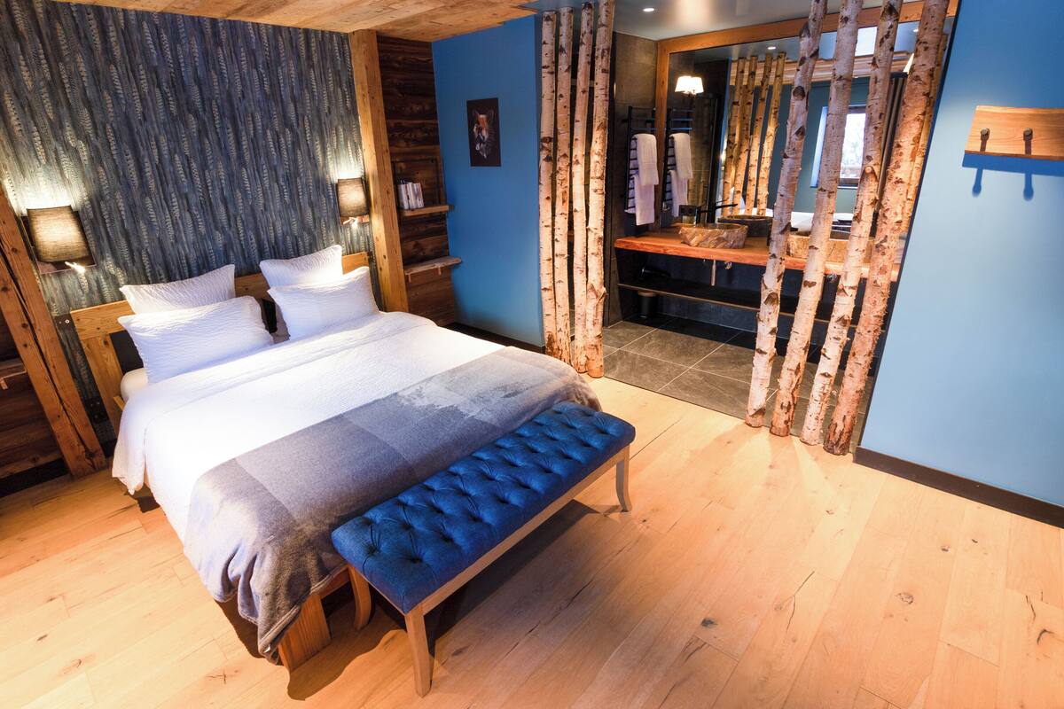 A bedroom with a large bed, a bench, wooden flooring, a blue wall, and a wooden headboard.