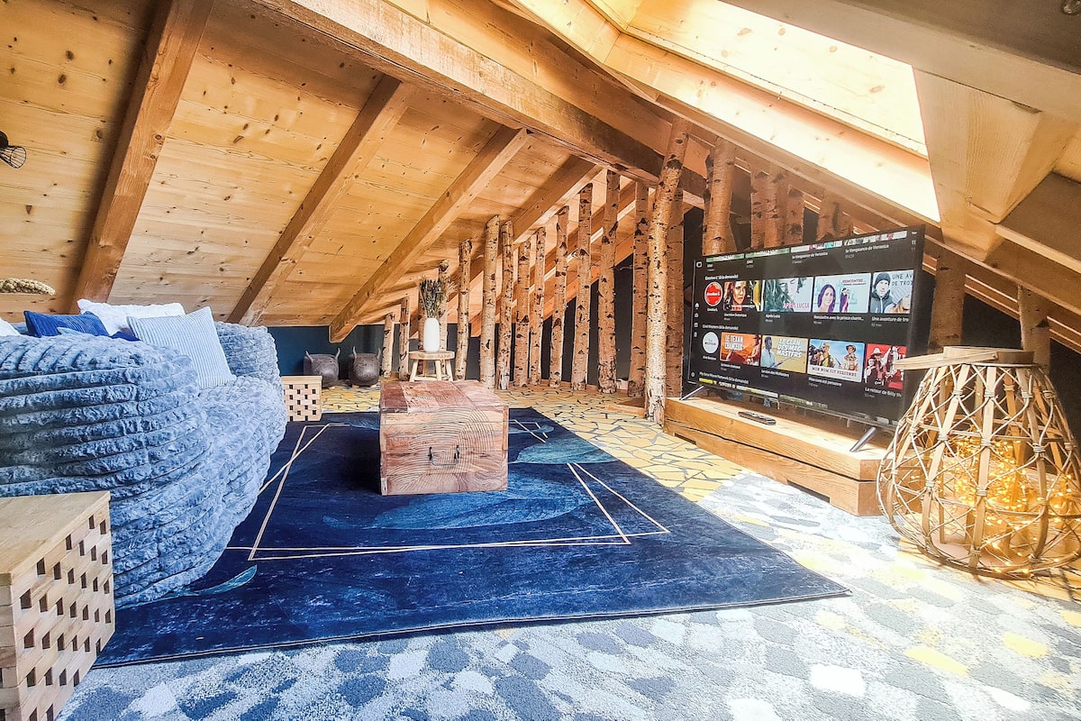 A cozy attic room with a wooden ceiling, a blue patterned rug, a wooden chest, a TV on the wall, and a decorative lamp.