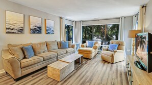 Ein geräumiges Wohnzimmer mit einem beigefarbenen Sofa, zwei Sesseln und einem Couchtisch. Große Fenster bieten Blick auf Bäume und einen Balkon.