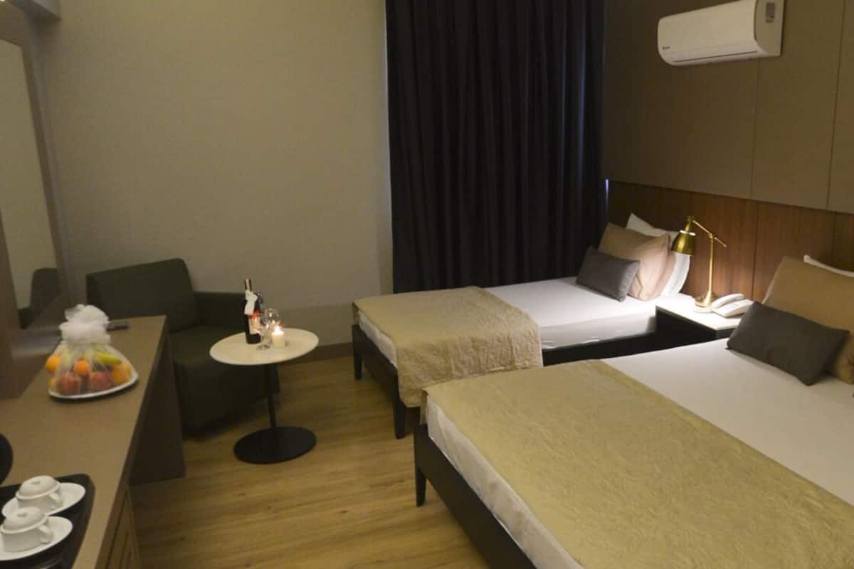 A hotel room with two beds, a small desk with a chair, a table with a fruit basket, and a wall-mounted air conditioner.