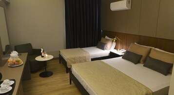 A hotel room with two beds, a small desk with a chair, a table with a fruit basket, and a wall-mounted air conditioner.