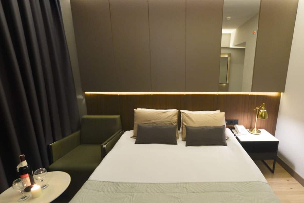 A hotel room with a bed, bedside table, lamp, and a small table with a bottle and glasses.