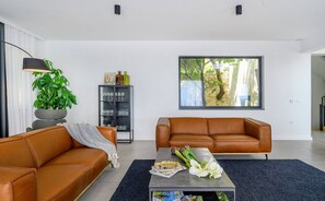 Uma sala de estar moderna com dois sofás de couro, uma mesa de centro, uma estante de livros e uma janela grande com vista para árvores.