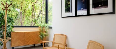 Une pièce avec une grande fenêtre, un meuble en bois, deux fauteuils en osier et des tableaux encadrés accrochés au mur.