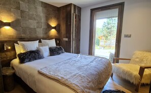 Quarto com uma cama grande, cabeceira de madeira, duas luminárias fixadas na parede, uma poltrona macia e uma porta de vidro deslizante para o exterior.