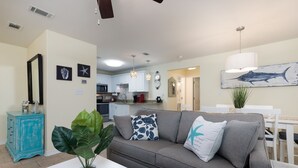 Un salon moderne avec un canapé gris, une table basse blanche et un meuble turquoise. On aperçoit une cuisine en arrière-plan, avec des meubles blancs et un luminaire suspendu.