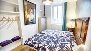 A bedroom with a bed, a dresser, a radiator, a window with curtains, a hanging light fixture, and a framed picture on the wall.
