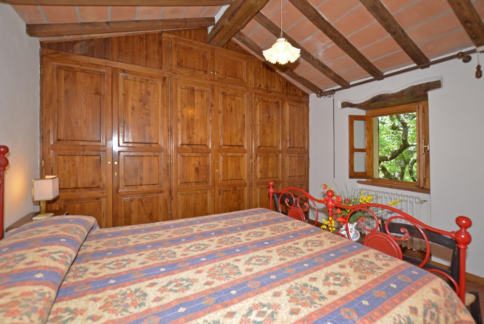 Una camera da letto con un'ampia armadiatura in legno, un letto con copriletto floreale e un poggiatesta in metallo rosso, e una finestra che si affaccia sul verde.
