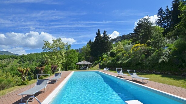 Ein rechteckiger Swimmingpool mit klarem, blauem Wasser, umgeben von einer Ziegelterrasse. Im Hintergrund sind Liegestühle und eine kleine Laube zu sehen.
