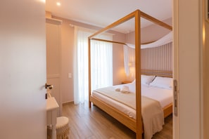 Ein Raum mit einem Himmelbett, Holzboden, Nachttisch und einem Fenster mit leichtem Vorhang.