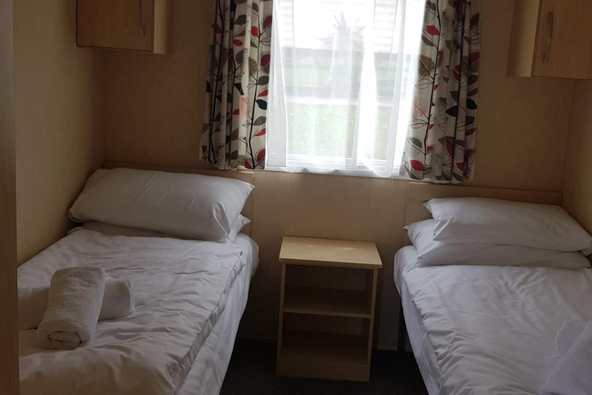 A room with two single beds, a small wooden nightstand between them, and a window with floral curtains.
