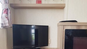 A flat-screen TV on a wooden stand in a room with a wooden shelf above holding wine glasses. - 2br-pool-garden-parking-wifi-sleeps6 (Paignton)