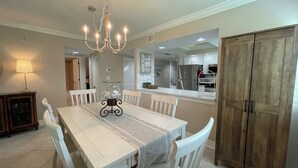 A dining area with a white table, chairs, a chandelier, and a wooden cabinet. - Essex 106N: 5 Min walk to Beach, Movies, Restaurants (Marco Island)