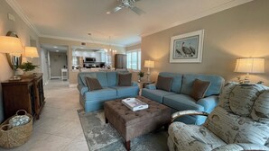 A spacious living room with a blue sofa set, a brown ottoman, and a patterned armchair. A kitchen is visible in the background. - Essex 106N: 5 Min walk to Beach, Movies, Restaurants (Marco Island)