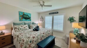 Un dormitorio con cama, un banco, una mesita de noche, una lámpara, un ventilador de techo y una ventana con persianas, con un letrero en la pared que dice 'BEACH HOUSE'.