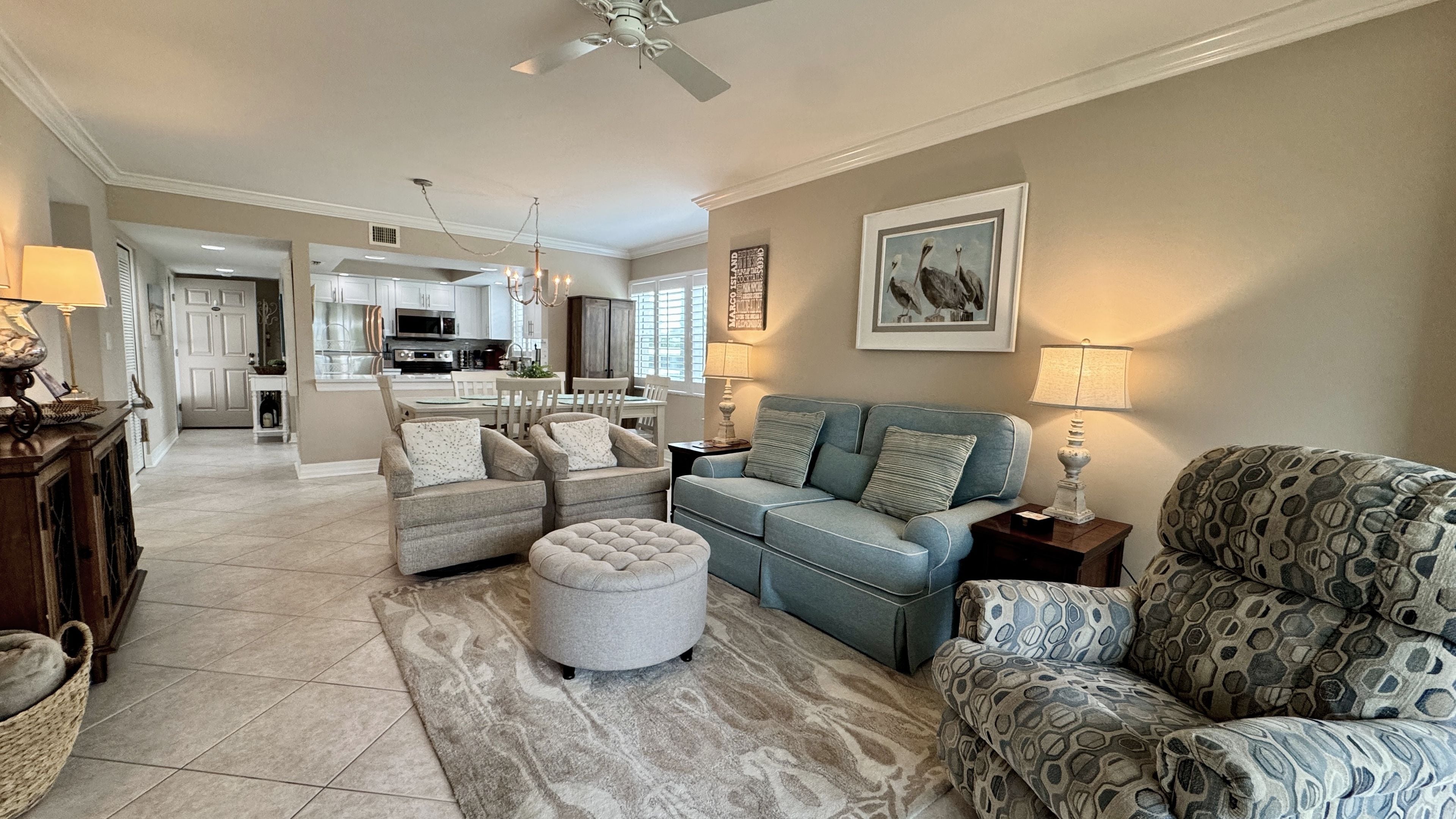 A spacious living room with a sofa set, ottoman, and patterned armchair, a dining area with a chandelier, and a kitchen in the background.