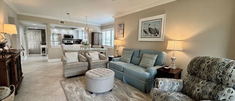 A spacious living room with a sofa set, ottoman, and patterned armchair, a dining area with a chandelier, and a kitchen in the background.