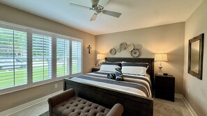 Une chambre à coucher comprenant un lit, une chaise, un ventilateur de plafond, une fenêtre avec des stores et un mur décoré d’assiettes.